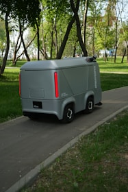 A small, futuristic-looking autonomous vehicle is traveling on a paved path through a lush park. The vehicle has a sleek gray exterior with red vertical lights on the rear. Tall trees with green foliage surround the path, and the ground is covered with grass and sparse vegetation.