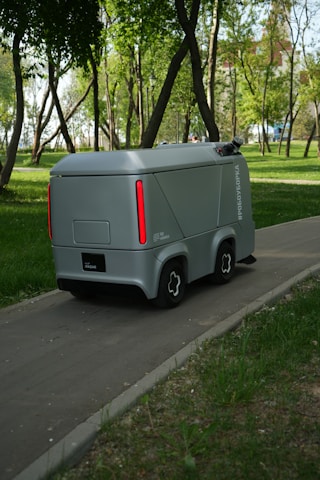 A small, futuristic-looking autonomous vehicle is traveling on a paved path through a lush park. The vehicle has a sleek gray exterior with red vertical lights on the rear. Tall trees with green foliage surround the path, and the ground is covered with grass and sparse vegetation.