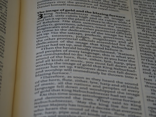 The image displays a close-up view of a page from a book, likely a religious text. The text is printed in a clear font with bold headings. Light shines on the page, casting shadows from the folds or binding of the book.