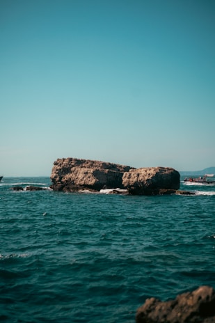 A rocky formation surrounded by deep blue ocean water under a clear sky. A small boat with a few people is visible near the rock formation, while distant land can be seen in the background.