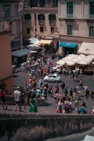 A busy outdoor plaza filled with people walking and gathering, surrounded by multi-story buildings with balconies. There are colorful storefronts with awnings, and a vintage white car is parked in the middle of the plaza. Numerous umbrellas provide shade for outdoor seating areas. The atmosphere is lively, with people of various attire suggesting casual summer wear.