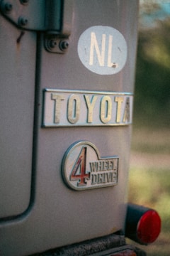 A close-up view of the rear of a vehicle, showcasing a Toyota emblem and a 4-wheel drive badge. There is also a circular sticker with the letters 'NL' on it. The focus is on the branding and identifiers, with a slightly vintage feel to the photograph.