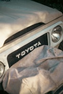 A close-up view of the front portion of a vintage Toyota vehicle. The image highlights the prominent grille with the Toyota logo in the center. There is a cloth draped over part of the grille and headlights, suggesting storage or protection.