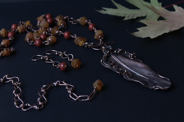 A handcrafted jewelry set featuring a necklace and matching earrings laid out on a dark surface. The necklace showcases a detailed feather pendant, surrounded by brown and reddish-brown beads and intricate wirework. The earrings mirror the aesthetic of the necklace with similar beadwork. A few dried, delicate leaves are present in the corner, adding a natural touch.
