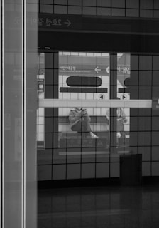 A black and white photo of a subway station, viewed through a glass panel. The image captures the reflection of a person sitting on a bench at the station, looking at a phone. Signage in Korean is visible in the background, highlighting the location and giving directions.