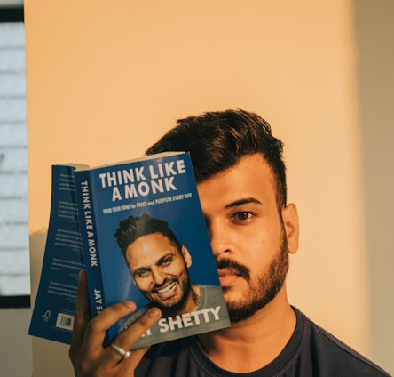 A man with a beard is holding up a book titled 'Think Like a Monk' by Jay Shetty. The book partially covers his face, showing a close-up portrait on the cover. The background is softly lit with a warm tone.
