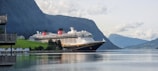 A large cruise ship is docked in a serene fjord with towering mountains in the background. The ship is white and black with red funnel accents, situated beside a green hillside with small cabins. The calm water reflects the scenery.