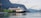 A large cruise ship is docked in a serene fjord with towering mountains in the background. The ship is white and black with red funnel accents, situated beside a green hillside with small cabins. The calm water reflects the scenery.