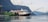 A large cruise ship is docked in a serene fjord with towering mountains in the background. The ship is white and black with red funnel accents, situated beside a green hillside with small cabins. The calm water reflects the scenery.