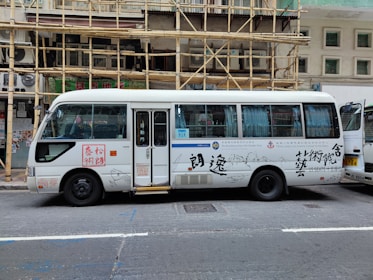 A white passenger minibus is parked on the street with Chinese calligraphy and art designs on the side. The minibus features a combination of blue and white curtains in the windows and is positioned next to a building with bamboo scaffolding. Several signs and text are visible on the bus, and a partially open door reveals the interior.