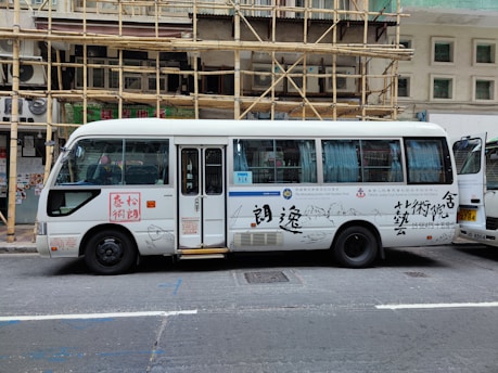 A white passenger minibus is parked on the street with Chinese calligraphy and art designs on the side. The minibus features a combination of blue and white curtains in the windows and is positioned next to a building with bamboo scaffolding. Several signs and text are visible on the bus, and a partially open door reveals the interior.