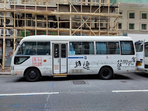 A white passenger minibus is parked on the street with Chinese calligraphy and art designs on the side. The minibus features a combination of blue and white curtains in the windows and is positioned next to a building with bamboo scaffolding. Several signs and text are visible on the bus, and a partially open door reveals the interior.