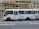 A white passenger minibus is parked on the street with Chinese calligraphy and art designs on the side. The minibus features a combination of blue and white curtains in the windows and is positioned next to a building with bamboo scaffolding. Several signs and text are visible on the bus, and a partially open door reveals the interior.