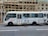 A white passenger minibus is parked on the street with Chinese calligraphy and art designs on the side. The minibus features a combination of blue and white curtains in the windows and is positioned next to a building with bamboo scaffolding. Several signs and text are visible on the bus, and a partially open door reveals the interior.