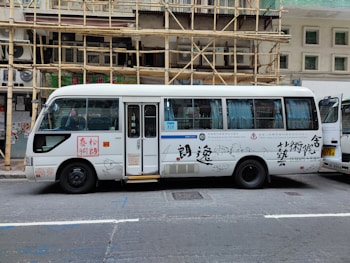 A white passenger minibus is parked on the street with Chinese calligraphy and art designs on the side. The minibus features a combination of blue and white curtains in the windows and is positioned next to a building with bamboo scaffolding. Several signs and text are visible on the bus, and a partially open door reveals the interior.