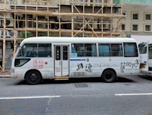 A white passenger minibus is parked on the street with Chinese calligraphy and art designs on the side. The minibus features a combination of blue and white curtains in the windows and is positioned next to a building with bamboo scaffolding. Several signs and text are visible on the bus, and a partially open door reveals the interior.
