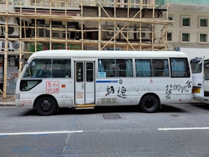 A white passenger minibus is parked on the street with Chinese calligraphy and art designs on the side. The minibus features a combination of blue and white curtains in the windows and is positioned next to a building with bamboo scaffolding. Several signs and text are visible on the bus, and a partially open door reveals the interior.