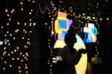 A silhouette of a person holding a camera, standing amidst a backdrop of colorful neon signs and strings of small lights wrapped around trees. The scene appears to be set at night, creating a contrast between the bright lights and the dark outlines of objects.