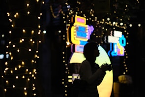 A silhouette of a person holding a camera, standing amidst a backdrop of colorful neon signs and strings of small lights wrapped around trees. The scene appears to be set at night, creating a contrast between the bright lights and the dark outlines of objects.