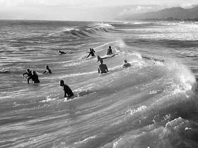 A group of surfers is captured riding the waves in the ocean. The surfers are scattered across the water, with surfboards visible beneath them. The surf creates a dynamic pattern of white foam against the darker ocean. Distant mountains and a shoreline can be seen on the horizon, adding depth to the scene.
