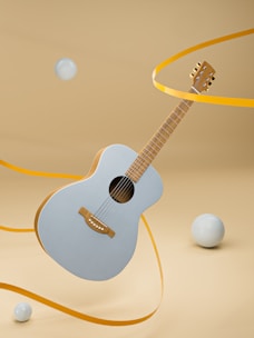 A light blue acoustic guitar is suspended in mid-air against a beige background. Yellow ribbons are winding around the guitar, and several light blue spheres are floating around it.