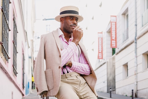 A person dressed in stylish clothing, including a beige blazer draped over one shoulder and a straw hat, stands against an urban street backdrop. The individual has a thoughtful expression and appears to be posing. The urban setting includes buildings with signs that read 'Les Abbesses', suggesting a street or neighborhood name.