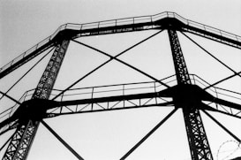 A black and white image of a large metal structure with intricate lattice patterns, likely an industrial framework or scaffolding. The structure is composed of interconnected beams and appears to extend upward into the sky.