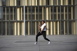A person is dancing energetically in an open area with a modern building as the backdrop. The individual is wearing a white shirt, black pants, and white sneakers. Long red braids flow with the movement, conveying motion and style.