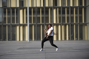 A person is dancing energetically in an open area with a modern building as the backdrop. The individual is wearing a white shirt, black pants, and white sneakers. Long red braids flow with the movement, conveying motion and style.