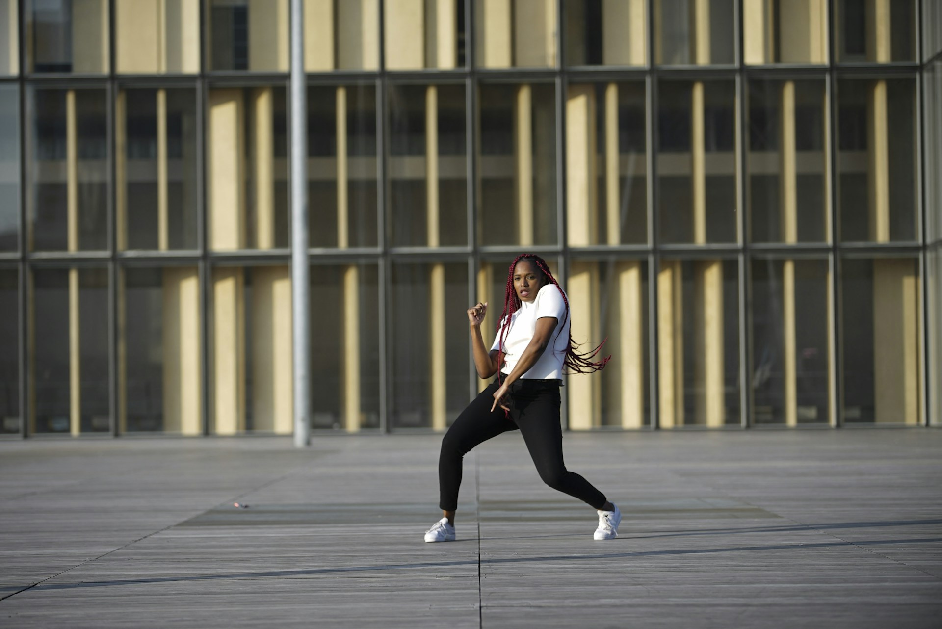 A person is dancing energetically in an open area with a modern building as the backdrop. The individual is wearing a white shirt, black pants, and white sneakers. Long red braids flow with the movement, conveying motion and style.