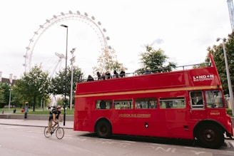 A bright red, double-decker London bus with an open top passes by, contrasting the greenery of trees and the iconic London Eye ferris wheel in the background. A man on a bicycle rides alongside the bus, adding a sense of movement to the scene filled with tourists and urban elements.
