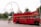 A bright red, double-decker London bus with an open top passes by, contrasting the greenery of trees and the iconic London Eye ferris wheel in the background. A man on a bicycle rides alongside the bus, adding a sense of movement to the scene filled with tourists and urban elements.