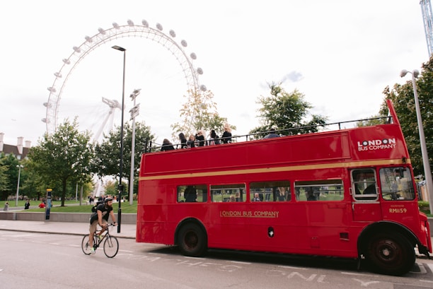 A bright red, double-decker London bus with an open top passes by, contrasting the greenery of trees and the iconic London Eye ferris wheel in the background. A man on a bicycle rides alongside the bus, adding a sense of movement to the scene filled with tourists and urban elements.