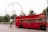 A bright red, double-decker London bus with an open top passes by, contrasting the greenery of trees and the iconic London Eye ferris wheel in the background. A man on a bicycle rides alongside the bus, adding a sense of movement to the scene filled with tourists and urban elements.