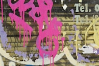 Vibrant pink spray paint drips over a weathered wall that has partial text reading 'Tel.' in faded yellow. Layers of peeling paint reveal several colors, including purple and white, adding texture to the surface.