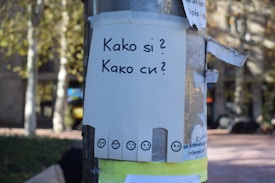 A public notice board displays a piece of grid paper with the text 'Kako si?' and 'Kako си?' written in bold letters. The paper is attached to a pole with old, torn layers of other paper and tape. At the bottom of the paper, there are several small squares each with an emoticon depicting different emotions. The background shows a blurred outdoor setting with trees and sunlight.
