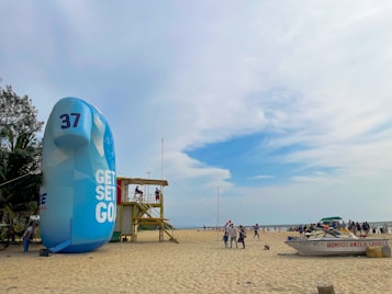A beach scene with a large inflatable structure displaying the text 'GET SET GO' and the number 37. Nearby, a wooden lifeguard tower is occupied by people. There are a few palm trees on the left and a boat labeled 'DOMROD WATER SPORTS' on the sand. The beach is populated by people enjoying the day under a partly cloudy blue sky.