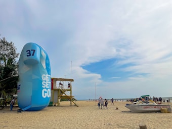A beach scene with a large inflatable structure displaying the text 'GET SET GO' and the number 37. Nearby, a wooden lifeguard tower is occupied by people. There are a few palm trees on the left and a boat labeled 'DOMROD WATER SPORTS' on the sand. The beach is populated by people enjoying the day under a partly cloudy blue sky.