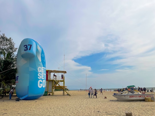 A beach scene with a large inflatable structure displaying the text 'GET SET GO' and the number 37. Nearby, a wooden lifeguard tower is occupied by people. There are a few palm trees on the left and a boat labeled 'DOMROD WATER SPORTS' on the sand. The beach is populated by people enjoying the day under a partly cloudy blue sky.