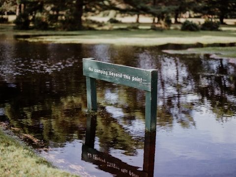 A wooden sign partially submerged in water reads 'No camping beyond this point.' The sign is surrounded by a flooded area with reflections of trees visible in the water, indicating recent heavy rainfall or high water levels. Lush greenery and trees are visible in the background, suggesting a park or natural setting.