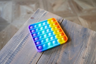 A multicolored pop-it fidget toy sits on a wooden table. The toy features a gradient of purple, blue, and yellow colors, forming a rectangular shape with bubble-like protrusions.