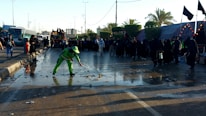 A person in bright green workwear is cleaning a wet street with a broom. There is a crowd of people dressed mostly in black gathered along the side of the road. The scene is set outdoors with palm trees and there are several buses and tents visible in the background.
