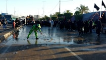 A person in bright green workwear is cleaning a wet street with a broom. There is a crowd of people dressed mostly in black gathered along the side of the road. The scene is set outdoors with palm trees and there are several buses and tents visible in the background.