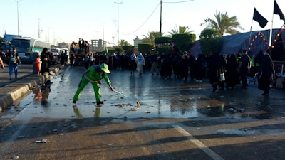 A person in bright green workwear is cleaning a wet street with a broom. There is a crowd of people dressed mostly in black gathered along the side of the road. The scene is set outdoors with palm trees and there are several buses and tents visible in the background.