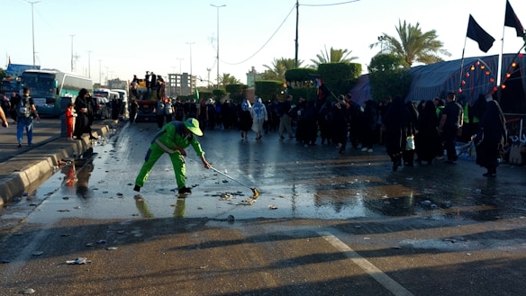 A person in bright green workwear is cleaning a wet street with a broom. There is a crowd of people dressed mostly in black gathered along the side of the road. The scene is set outdoors with palm trees and there are several buses and tents visible in the background.