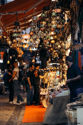 A bustling market street filled with colorful hanging lanterns. People are browsing through various stalls displaying intricately designed lamps and decorative items. The atmosphere is vibrant and busy, indicating a popular shopping area.