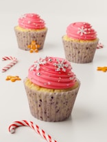 Three cupcakes topped with swirls of pink frosting decorated with snowflakes and silver beads are arranged on a light background. Gingerbread man cookies and candy canes are scattered around, adding a festive touch.