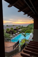A scenic view of a resort with a large, rectangular swimming pool surrounded by lounge chairs and green palm trees. The setting includes a multi-story building with wooden railings and a sunset in the background, casting a warm glow over the ocean and distant hills.