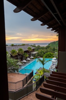 A scenic view of a resort with a large, rectangular swimming pool surrounded by lounge chairs and green palm trees. The setting includes a multi-story building with wooden railings and a sunset in the background, casting a warm glow over the ocean and distant hills.