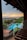A scenic view of a resort with a large, rectangular swimming pool surrounded by lounge chairs and green palm trees. The setting includes a multi-story building with wooden railings and a sunset in the background, casting a warm glow over the ocean and distant hills.